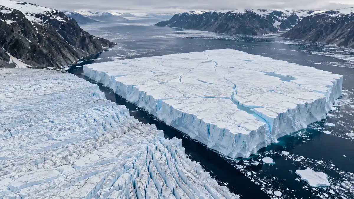 Vista aérea de una isla de hielo tabular separada del frente del glaciar por un canal de agua oscura, en un fiordo ártico con laderas rocosas nevadas
